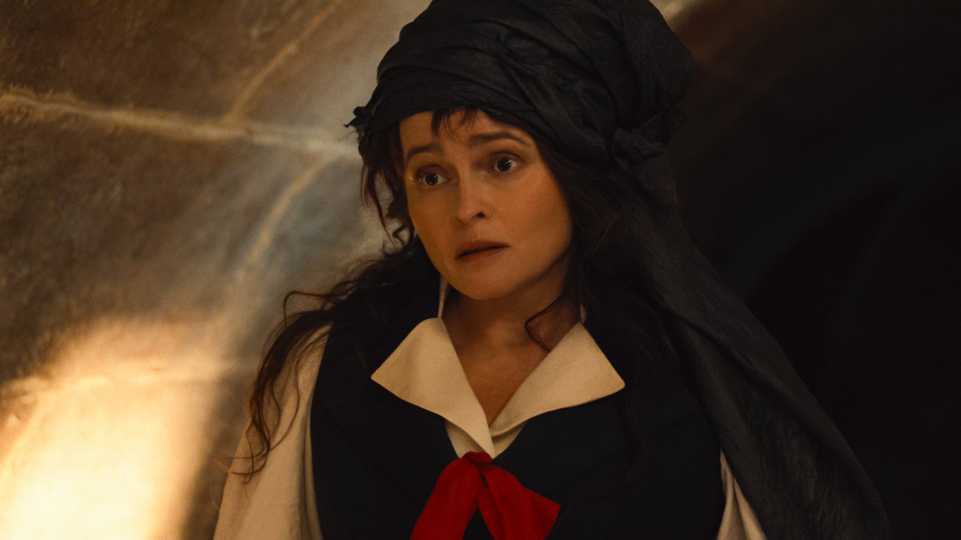 "Woman dressed in period costume with a black headdress and red ribbon stands in a dimly lit stone hallway, looking surprised or concerned. Warm light highlights the stone wall in the background."