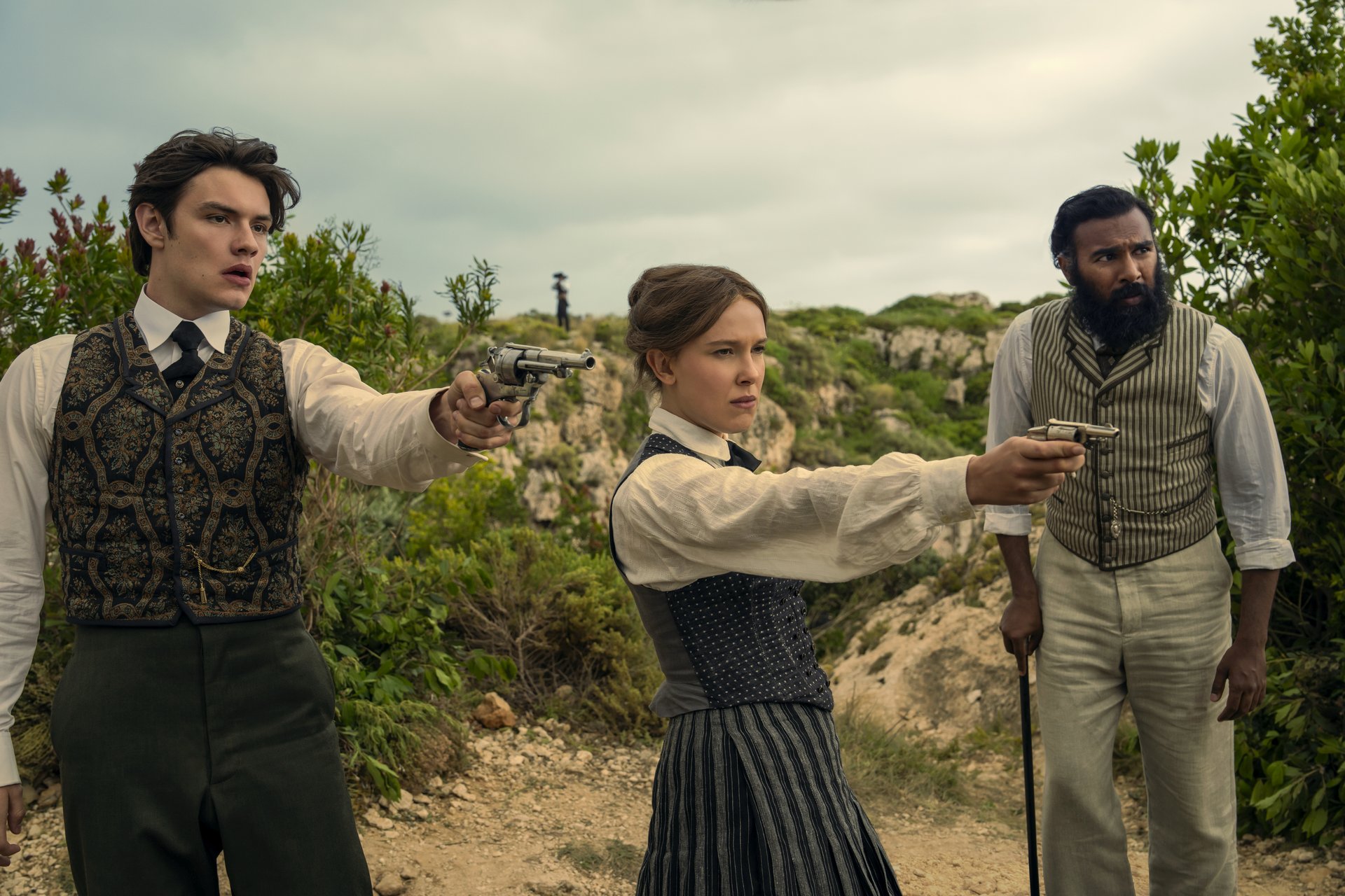 "Three people in vintage clothing stand outdoors in a rugged, green landscape; two point pistols ahead while the third stands beside them with a cane, all looking alert and tense."