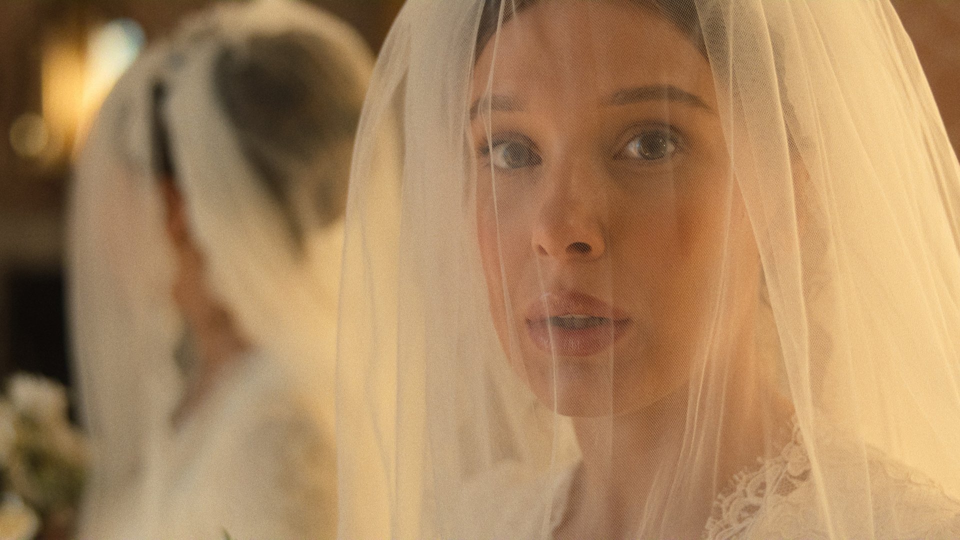 "A bride with a white veil and lace dress stands indoors, looking ahead with a soft expression. A mirror reflects her image, and flowers are visible in the background, creating an elegant, warm atmosphere."