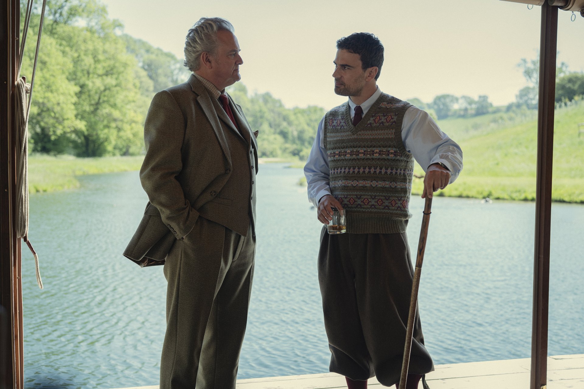 "Two men in aristocratic clothing talk by a riverbank; one holds a drink, the other a walking stick. They stand under a shaded structure with green trees and calm water visible in the background on a sunny day."