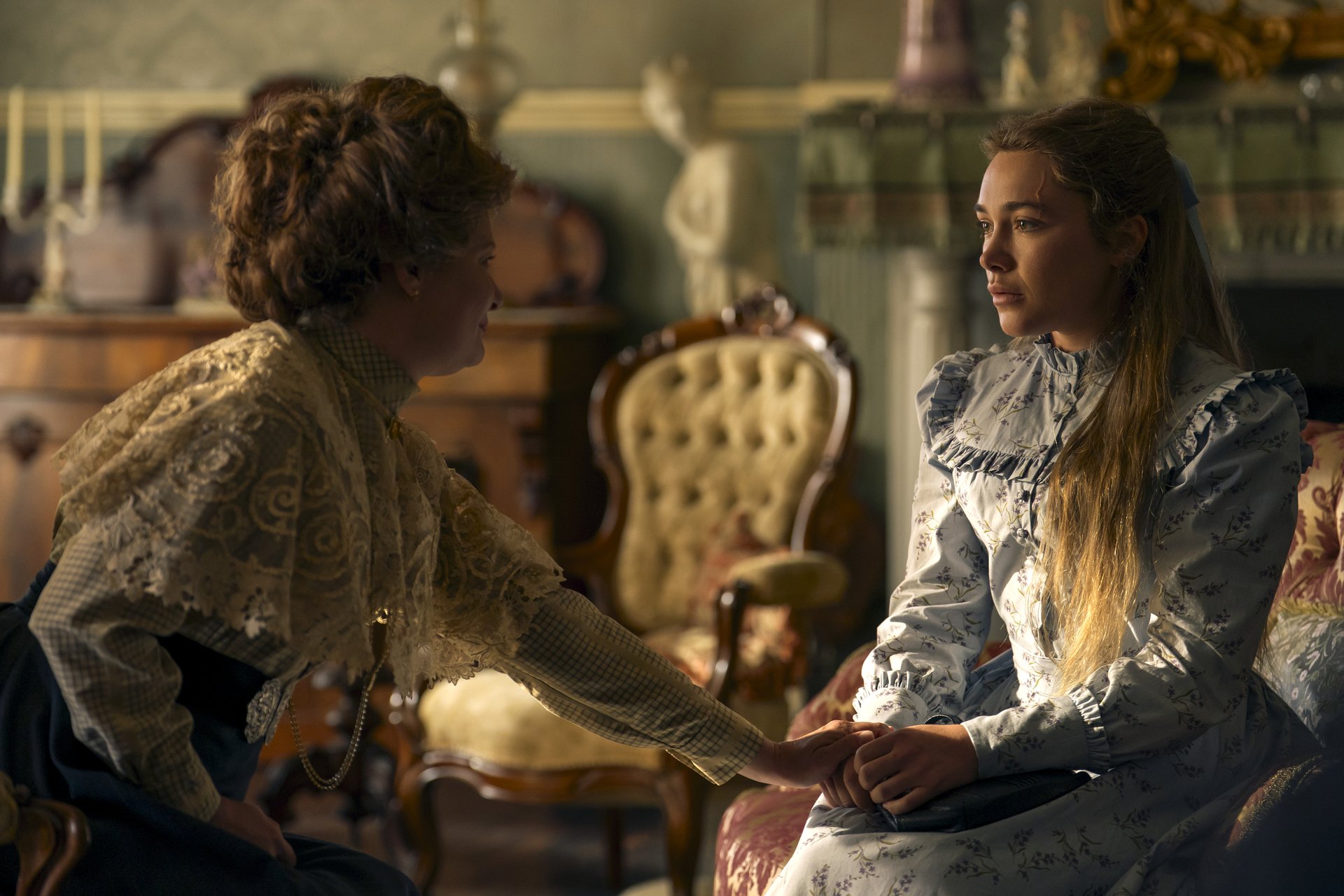 "Two women in Victorian-era dresses sit in an ornate, vintage living room with antique chairs and decor; one woman gently touches the other's hand, suggesting a comforting or emotional moment."