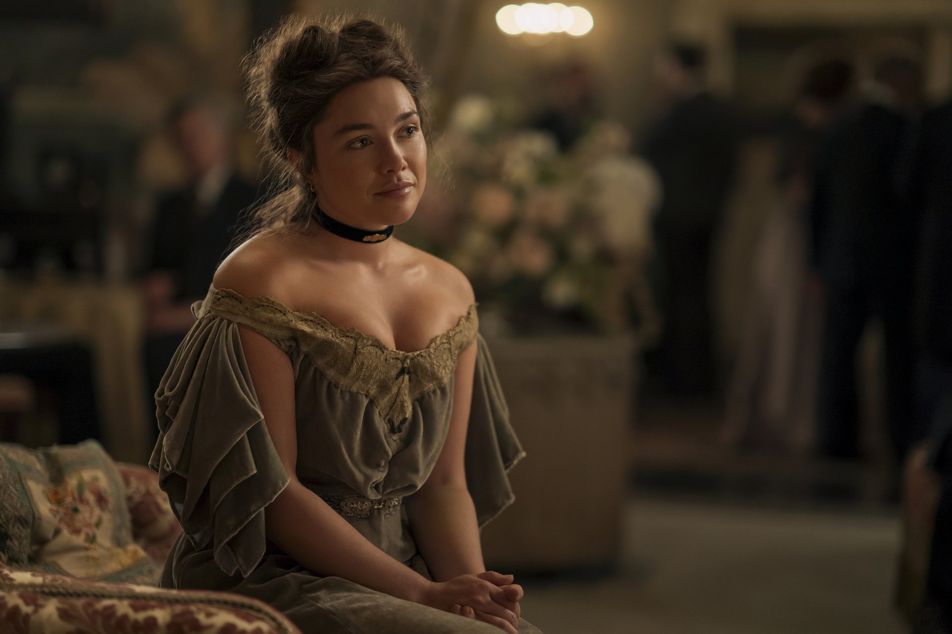 "A woman in a vintage off-shoulder dress sits on a patterned sofa in a softly-lit, elegant room with blurred people and floral arrangements in the background, suggesting a formal or historic gathering."