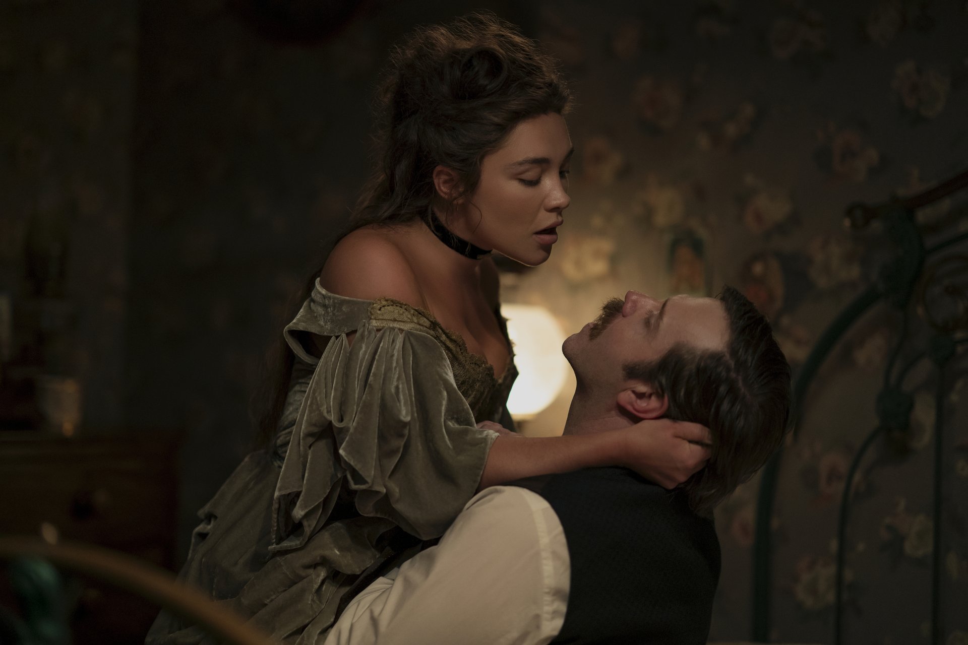 "A woman in period costume sits on a man's lap in an intimate embrace in a dimly lit, vintage bedroom with floral wallpaper and antique furniture."