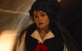 "Woman dressed in period costume with a black headdress and red ribbon stands in a dimly lit stone hallway, looking surprised or concerned. Warm light highlights the stone wall in the background."
