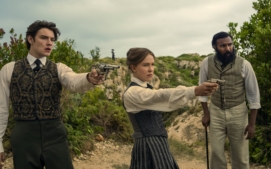 "Three people in vintage clothing stand outdoors in a rugged, green landscape; two point pistols ahead while the third stands beside them with a cane, all looking alert and tense."