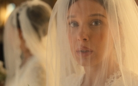"A bride with a white veil and lace dress stands indoors, looking ahead with a soft expression. A mirror reflects her image, and flowers are visible in the background, creating an elegant, warm atmosphere."