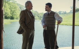"Two men in aristocratic clothing talk by a riverbank; one holds a drink, the other a walking stick. They stand under a shaded structure with green trees and calm water visible in the background on a sunny day."