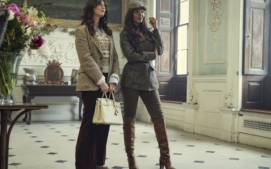 "Two stylish women stand in an elegant, classic interior with tall windows and ornate decor. One holds a white handbag, both wear blazers and boots, appearing poised and fashionable in a well-lit, sophisticated room with flowers and art."