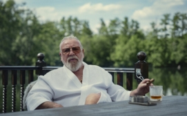 "Older man in white robe sits outdoors by a wooden table, holding a cigar and drink, with greenery and a lake in the background, under a partly cloudy sky."