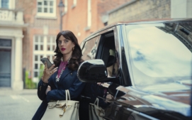 "Woman with long dark hair in a stylish blazer stands next to a black luxury car on a city street, holding a smartphone and beige purse, with upscale buildings in the background."