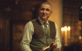 "Middle-aged man in a checkered shirt and vest holding a book, standing indoors with warm lighting, blurred background features candles and wooden decor, creating a cozy, intimate atmosphere."