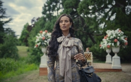 "Woman with long dark hair in an elegant outfit and bow scarf stands outdoors in a lush garden with large floral arrangements and greenery, holding a black handbag on her arm."