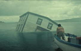 "A partially submerged house tilts in a misty lake as three people in a small boat observe, with cloudy skies and distant hills creating a dramatic, mysterious atmosphere."