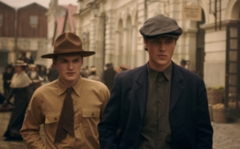 "Two young men in vintage clothing and hats walk on an old city street with historical buildings and people in period attire in the background, evoking an early 20th-century urban setting."