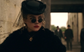 "Woman in vintage black attire with round sunglasses, a veiled hat, and gold jewelry stands near a stone wall in an urban setting, with blurred people and old-style architecture in the background."