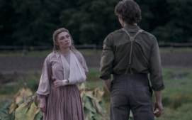 "A woman with her arm in a sling faces a man in suspenders outdoors, standing near large green leaves in a rural farm setting with trees and a field in the background."
