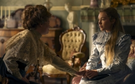 "Two women in Victorian-era dresses sit in an ornate, vintage living room with antique chairs and decor; one woman gently touches the other's hand, suggesting a comforting or emotional moment."