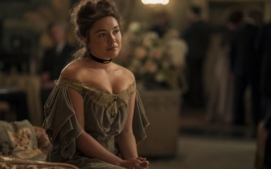 "A woman in a vintage off-shoulder dress sits on a patterned sofa in a softly-lit, elegant room with blurred people and floral arrangements in the background, suggesting a formal or historic gathering."
