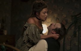 "A woman in period costume sits on a man's lap in an intimate embrace in a dimly lit, vintage bedroom with floral wallpaper and antique furniture."